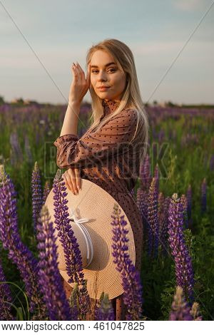 A Beautiful Woman In A Straw Hat Walks In A Field With Purple Flowers. A Walk In Nature In The Lupin