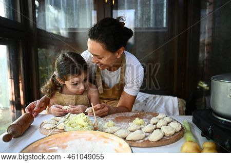 Loving Mother Hugging Her Adorable Little Daughter, Standing Together At A Table With Dumplings On A