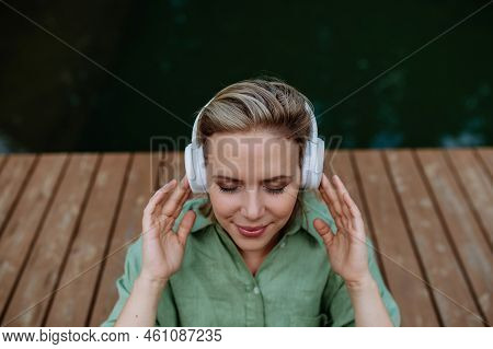 Rlaxed Woman Wearing Headphones Listening To Music Sitting On A Pier By Natureal Lake In Summer