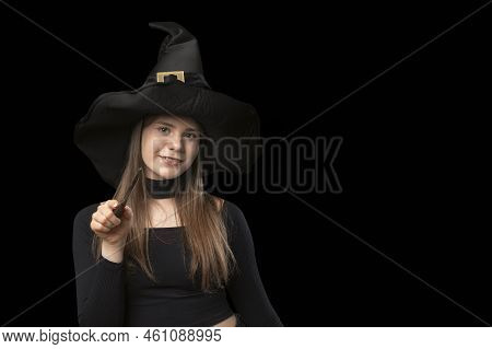 Porttrait Of Cute Young Witch In Pointed Hat With Magic Wand In Her Hands. Isolated On Black Backgro