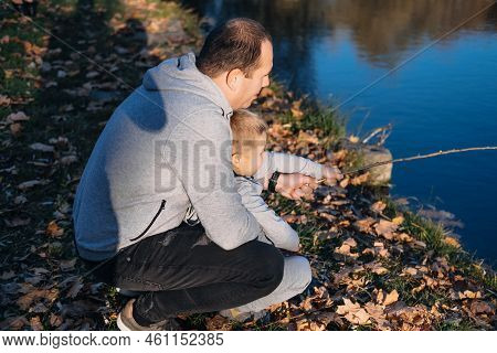 Father And His Son Sitting In Front Of The Lake Which Is Full Of Fishes Imitating Fishing By Holding