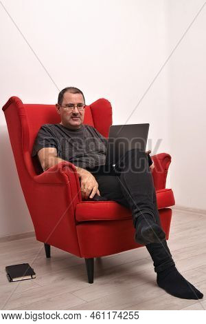 Middle-aged Man With A Laptop On His Lap Sed In A Red Armchair. The Man Is Holding Two Smartphones