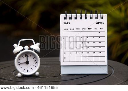 Alarm Clock Pointing At 9 O'clock With April 2023 Calendar On Blurred Park Background. Time And Cale