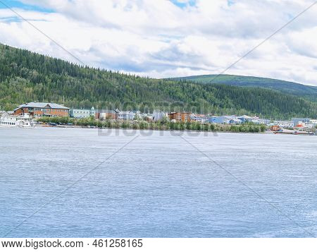 Dawson City Across The Yukon River With Rolling Tree Clad Hill Background.