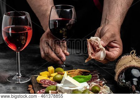 Hands Holding A Glass Of Wine And A Wooden Board With Different Kinds Of Cheese And Ham, Prosciutto,