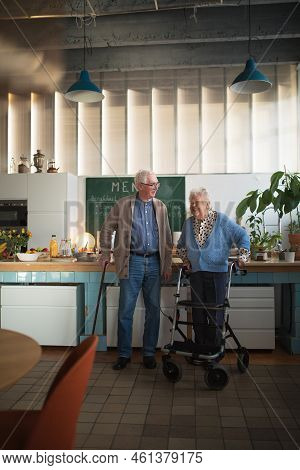 Senior Man And Woman Walking With Walker Indoors In Retirement Nursing Hme.