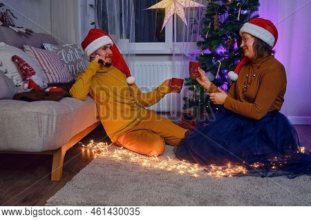 An Adult Man And A Woman Drink From A Cup At The Christmas Tree. Home Family Comfort On New Year Eve
