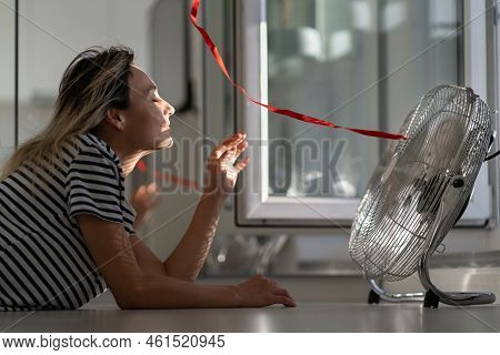 Smiling Young Woman Lying On The Floor In Front Of Working Electric Fan In Sunny Hot Summer Day, Enj