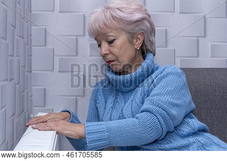 Elderly Woman 60-65 Years Old In A Warm Sweater Warming Near The Heating Radiator. Concept: Low Room