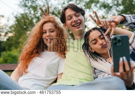 Three Young Smiling Friends Sitting On A Park Bench Taking Self-portrait On Smartphones. Concept Of 