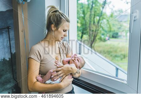 Caucasian Woman With Blond Hair Holding Her Newborn Baby Boy In Her Arms Smiling Standing By The Win
