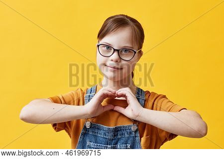 Waist Up Portrait Of Teen Girl With Down Syndrome Looking At Camera And Showing Heart Sign While Sta