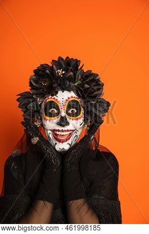 Excited Woman In Spooky Halloween Makeup And Black Wreath Holding Hands Near Face And Looking At Cam