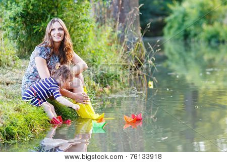 Adorable Little Girl And Her Mom Playing With Paper Boats In A River.