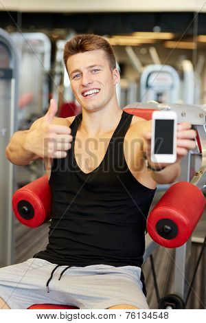 sport, bodybuilding, lifestyle, technology and people concept - smiling young man showing smartphone and thumbs up gesture in gym