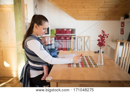 Beautiful young mother at home with her baby son sleeping in sling, reading a book or studying something