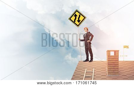 Young determined businessman standing on house roof and showing yellow roadsign. Mixed media