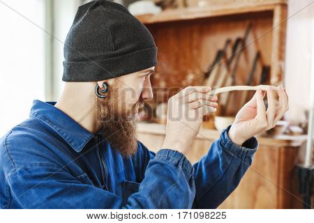 Worker with a beard wearing blue jeans suit and black hat holding and looking at wooden spoon, woodcarving, portrait, copy space.