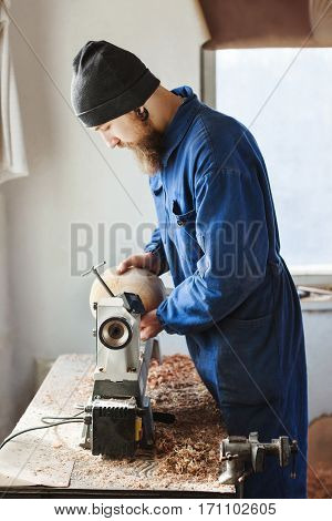 Worker with a beard wearing blue jeans suit and black hat working near table, woodcarving instruments on table, big window at background, portrait, copy space.