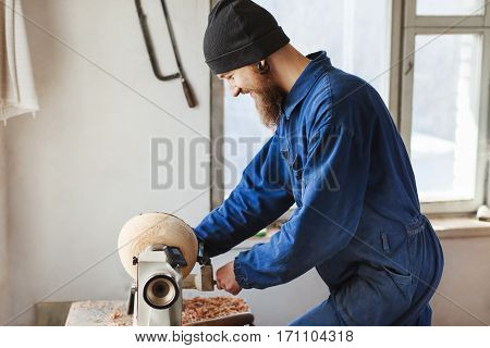 Worker with a beard wearing blue jeans suit and black hat working near table, woodcarving instruments on table, window at background, portrait, woodworking.