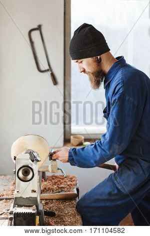 Man with a beard wearing blue jeans suit and black hat working near table, woodcarving instruments on table and on wall, window at background.
