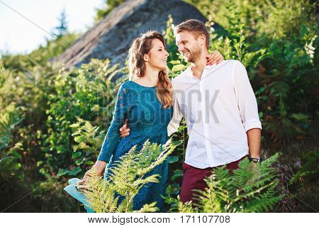 Nice couple standing together among fern and trees, outdoor, in the countryside. Beloved looking at each other, embracing each other by one hand and smiling. Woman wearing blue dress and has pink manicure and man wearing white shirt, claret trousers.