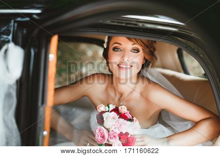 Wedding day. Bride sitting in wedding car, looking at camera, smiling and holding bouquet. Decorations. Waist up