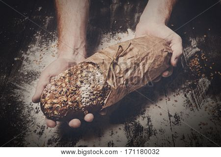 Warm fresh rye whole grain bread, wrapped in craft paper. Healthy baking and cooking concept background. Hands hold loaf on wooden table background, sprinkled with flour. Soft toning, filtered
