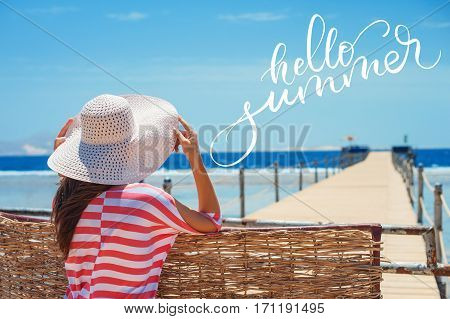 Closeup back view of woman in white hat looking out towards blue ocean and sky and words Hello summer. Calligraphy, lettering.