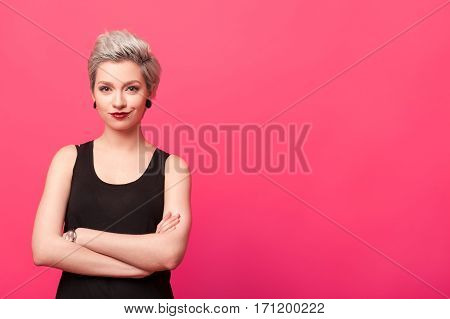 Close-up Portrait of young confident woman standing with arms crossed isolated over pink color background. Dyed blond short hairstyle