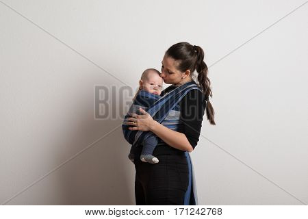 Beautiful young mother with her baby son in sling, kissing him on head. Studio shot on white background.