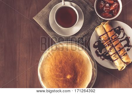 Stack of delicious homemade pancakes or blini with chocolate, cup of tea and jam in a bowl on wooden table. Toned photo. Morning healthy breakfast. Top view.