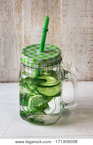 Mason jar mug with handle, lid and straw with detox cucumber agua fresca water with lime and mint ingredients, on rustic wood kitchen table, spring cleansing
