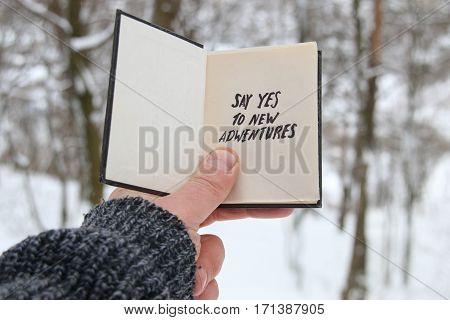 Hand holding a book with the inscription Say yes to new adventures on the background of the forest with snow-covered trees