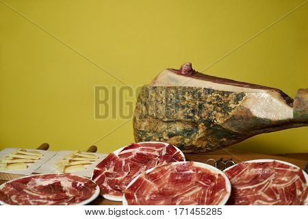 Smoked leg of lamb slised on white plate and cheese slices on wooden board with yellow background with copy space.