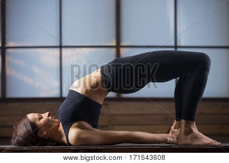 Young attractive woman practicing yoga, stretching in Glute Bridge exercise, dvi pada pithasana pose, working out, wearing sportswear, black tank top, pants, indoor full length, evening practice