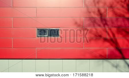 The front facade of a modern building of gray and red panels wih small window. Shadows of the trees on the facade of the building. Architectural background.