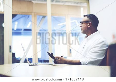 Handsome skilled wondered businessman sitting at his office desk and looking thoughtful. Ambitious pensive entrepreneur holding notebook after viewing laptop monthly report of the company's revenues