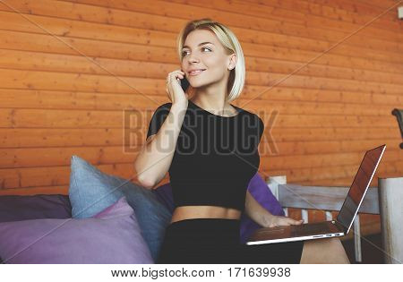 Young woman is smiling for friends during talk via smart phone while is sitting in modern co-working space.Beautiful hipster girl is calling via cell telephone during work on portable laptop computer