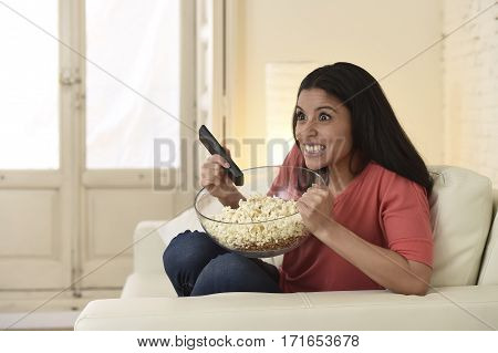 young beautiful Spanish woman in jeans home alone watching television laughing sitting at sofa couch on living room happy and excited enjoying comedy movie having fun holding remote