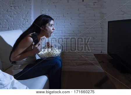 young beautiful latin woman home alone watching television smiling sitting at sofa couch on living room happy and excited enjoying TV show or movie relaxed in dark light eating popcorn
