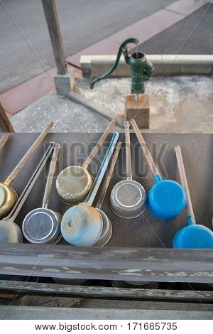 Multiple metal and plastic ladles on a table and a hand water pump next to a graveyard. Hand ladles are used as a part of Shinto ceremonies. Nobeoka Japan.