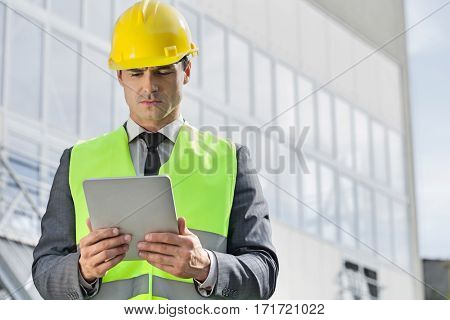 Young male engineer in reflector-vest and hardhat using digital tablet outside industry
