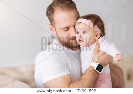 My adorable child. Happy peaceful young father sitting on the couch at home and hugging his newborn while expressing care and love