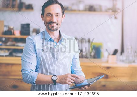 Happy to see you. Positive handsome cafe owner holding tablet and wearing apron while welcoming guests