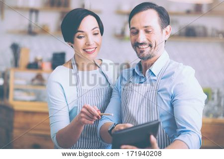 Express your ideas. Cheerful positive senior couple using tablet and discussing ideas while running the cafe