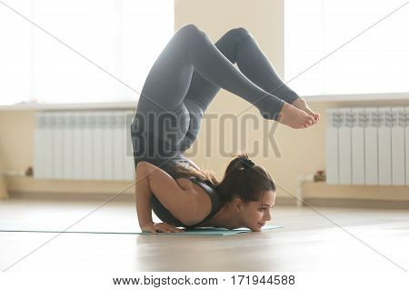 Young healthy attractive woman practicing yoga, standing in Ganda Bherundasana exercise, Formidable Face pose, working out, wearing grey sportswear, full length, beautiful home interior