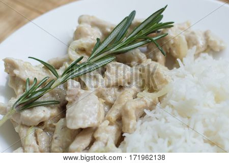 Portion of sliced chicken breast cooked in white sauce and fresh rosemary and served on a white plate with rice