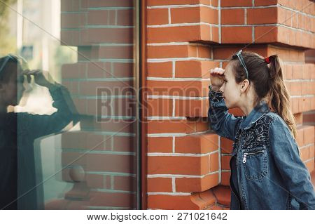 Teenage Girl In Blue Denim Dress Look In Window Of Building From Red (brown) Brick Outdoors. Young S