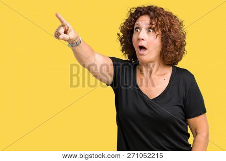 Beautiful middle ager senior woman over isolated background Pointing with finger surprised ahead, open mouth amazed expression, something in front